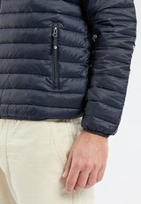 Veste matelassée bleu marine avec une poche zippée sur la poitrine gauche, en matériau nylon, poignets côtelés et design léger.