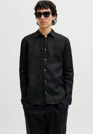 Férfi fekete hosszú ujjú, gombos ingben és fekete nadrágban, zsebbe dugott kézzel, nagy fekete napszemüveget viselve áll.