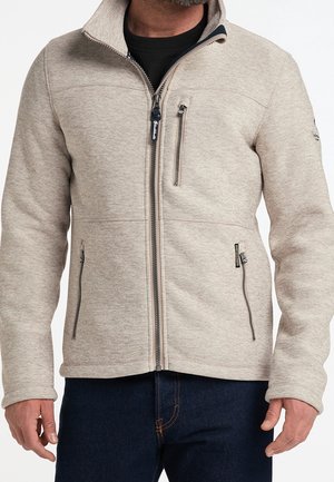 Homme portant une veste beige zippée avec trois poches zippées et un jean bleu foncé, debout, les mains détendues le long du corps.