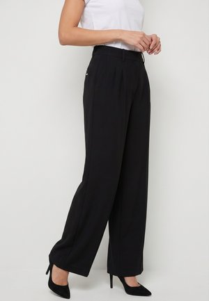 Femme portant un pantalon noir taille haute à jambes larges, un haut blanc ajusté à manches courtes et des talons hauts noirs à bout pointu, debout de profil.