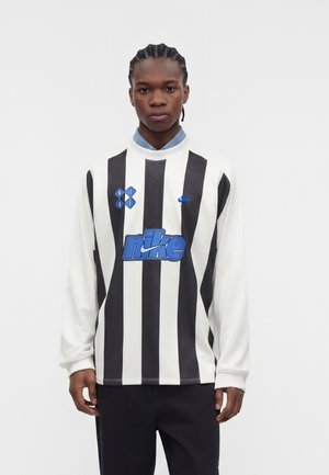 Mężczyzna w czarno-białej pionowo paskowanej bluzie Nike z niebieskimi logo, stojący na jednolitym białym tle.
