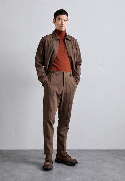 Bruine ritsjack, geribbelde oranje coltrui, bijpassende bruine broek en donkerbruine suède laarzen. Minimalistisch ontwerp met elastische tailleband.