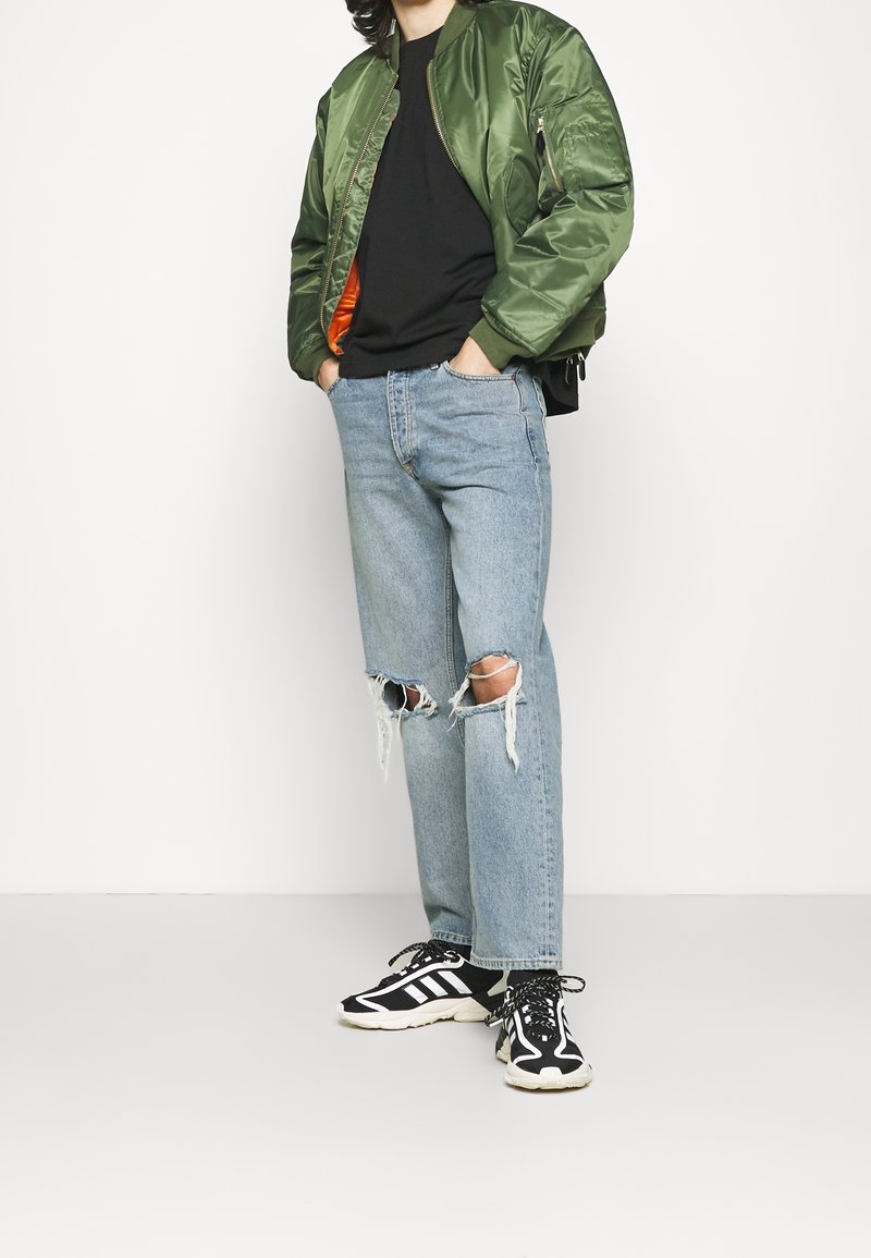 Zelená bomber bunda s žebrovanými manžetami, černé tričko, světle modré roztrhané džínsy a černobílé tenisky s texturovanou podrážkou.