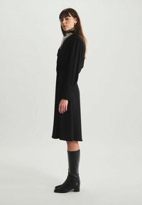 Robe noire à manches longues, taille cintrée et jupe évasée. Portée avec des bottes en cuir noires jusqu'aux genoux et une écharpe à motifs autour du cou.