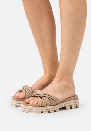 Chanclas de goma nude con un diseño de tiras retorcidas, suela gruesa y texturizada, y patrón recortado. Estilo cómodo para poner y quitar con una base robusta.