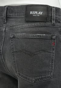 Juodi džinsai iš džinso su odine etikete ant juosmens, pažymėta "REPLAY" ženklu, ir raudonai siuvinėtu logotipu ant galinės kišenės.