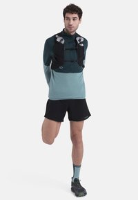 Chaleco de hidratación negro con acentos grises sobre una camiseta de manga larga color teal, combinado con pantalones cortos negros y zapatillas de trail verdes. Calcetines a rayas teal.