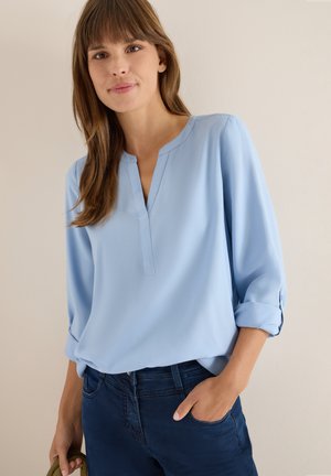 Blusa azzurro chiaro a maniche lunghe con scollatura a V e polsini arrotolati; tessuto morbido; abbinata a jeans blu scuro e una borsa verde.