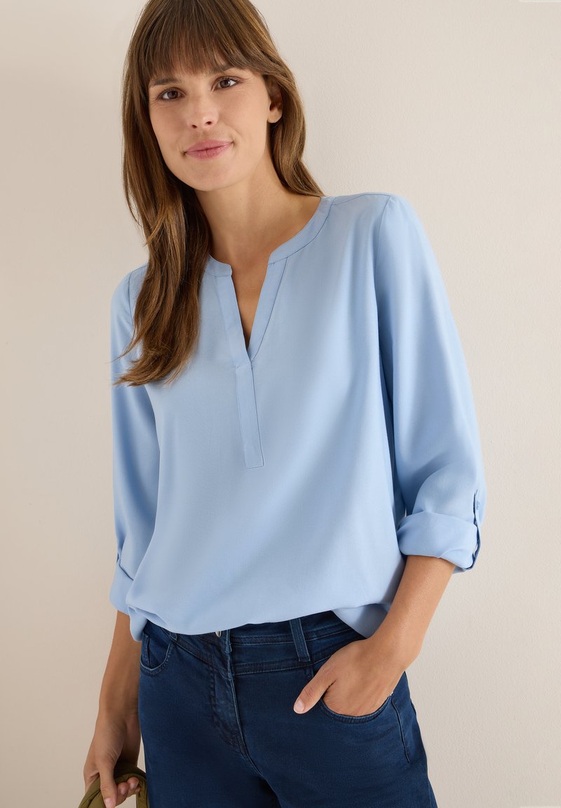 Lyseblå, langærmet bluse med V-udskæring og opsmøgede manchetter; glat stof; parret med mørkeblå jeans og en grøn taske.