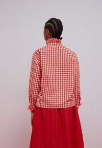 Red and white gingham-patterned blouse with a high collar, long sleeves, and gathered cuffs, paired with a solid red textured skirt.
