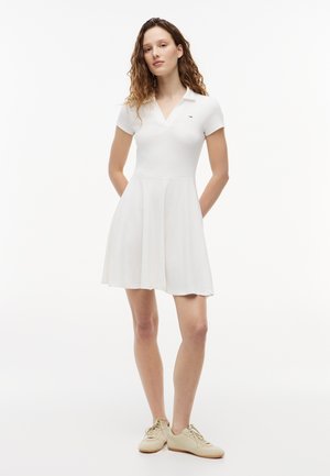 Frau steht mit verschränkten Händen hinter dem Rücken, trägt ein weißes Kurzarm-Polokleid und beige Sneakers vor weißem Hintergrund.