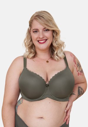 Lächelnde Frau mit blonden Haaren, die einen olivgrünen BH und passende Unterwäsche trägt und Arm- sowie Seitentattoos zeigt, vor weißem Hintergrund.