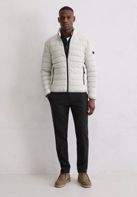 Jasnoszara kurtka puchowa z poziomym pikowaniem, kieszeniami na suwak i stójką, w połączeniu z czarnymi spodniami i beżowym obuwiem.