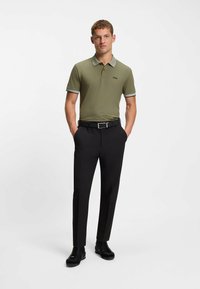 Polo verde oliva con un cuello estampado y detalles negros, combinado con pantalones negros y zapatos negros. Diseño sencillo y ajustado.
