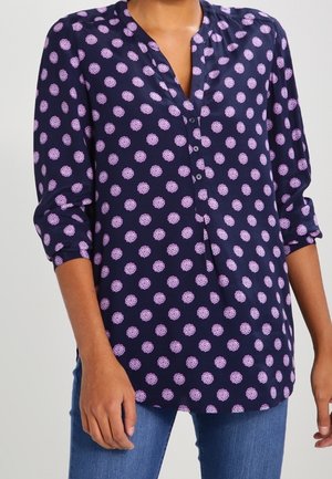 Woman wearing a navy blue blouse with pink circular floral patterns and three-quarter sleeves, paired with blue jeans.