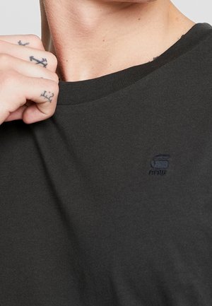 Close-up of person wearing dark gray T-shirt with small embroidered logo on chest, holding collar with tattooed hand.