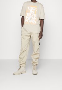 Beige short-sleeve T-shirt with white and orange graphic text, paired with loose beige cargo pants and light beige boots.
