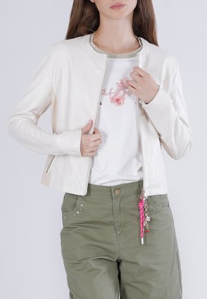 Nainen yllään valkoinen vetoketjullinen takki kukkakuvioisen valkoisen t-paidan päällä, oliivinvihreät housut, joissa on tähti- koristeluja, sekä avaimenperäkoru.