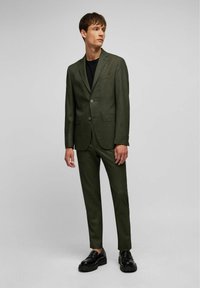 Costume sur mesure vert foncé avec un motif texturé, veste à un bouton et pantalon slim-fit. Porté avec une chemise noire et des chaussures noires.