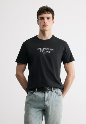 Mladý muž má na sebe čierne tričko Calvin Klein s bielym nápisom "CALVIN KLEIN EST. 1968" zastrčené do svetlomodrých džínsov s čiernym opaskom.