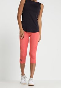 Mustat hihattomat urheilupaidat yhdistettynä kirkkaan korallinvärisiin capri-legginseihin, joissa on viistoja yksityiskohtia ja tekstilogo. Kengät täydentävät asua.