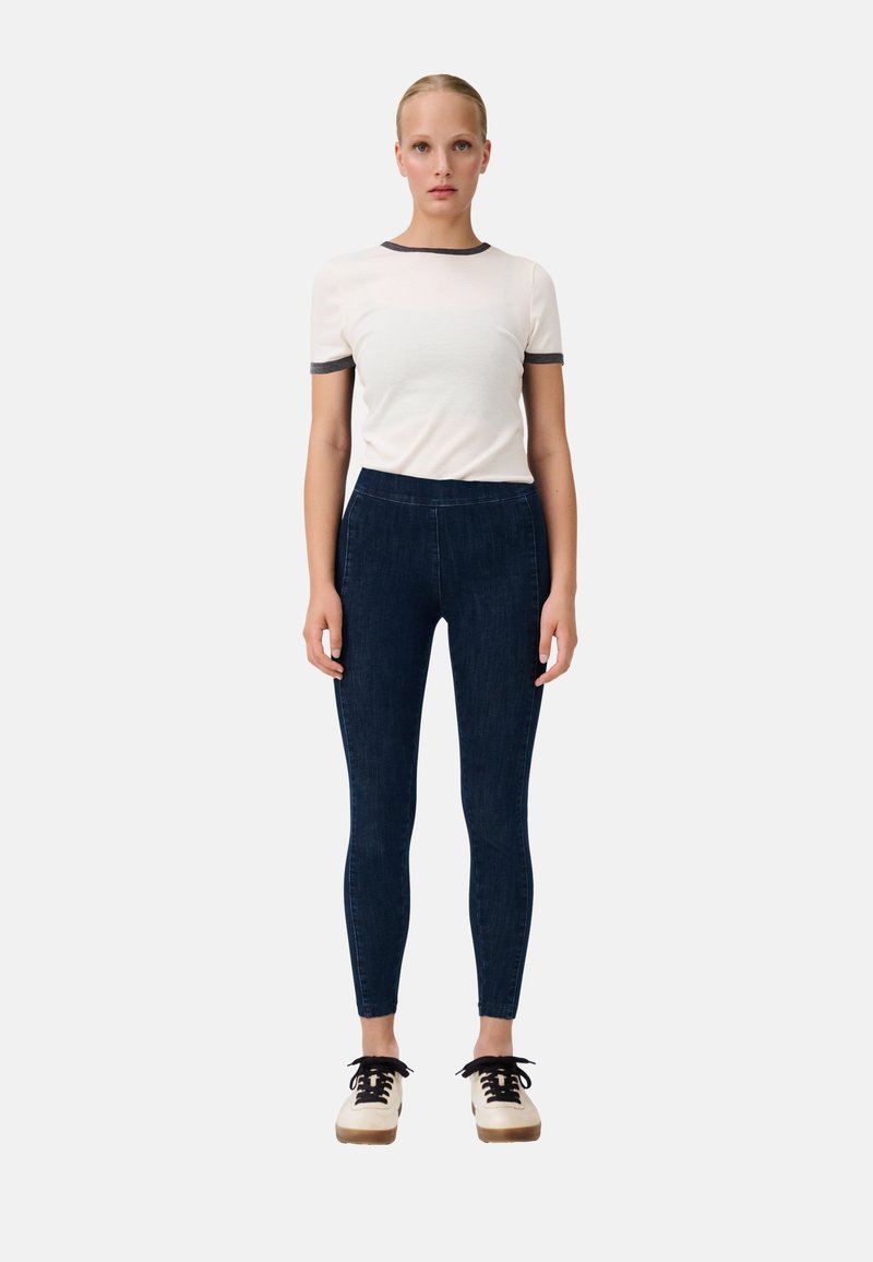 Weißes Kurzarm-Oberteil mit dunkelgrauem Saum, kombiniert mit dunkelblauen Skinny-Jeans und beigen Plateauschuhen mit schwarzen Schnürsenkeln.