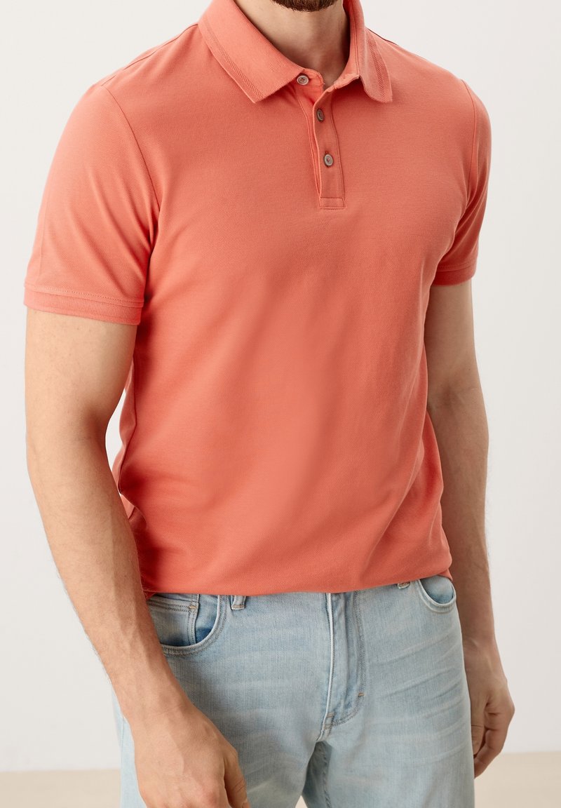 Homme portant un polo corail à manches courtes et un jean bleu clair, debout dans une posture détendue devant un fond uni.