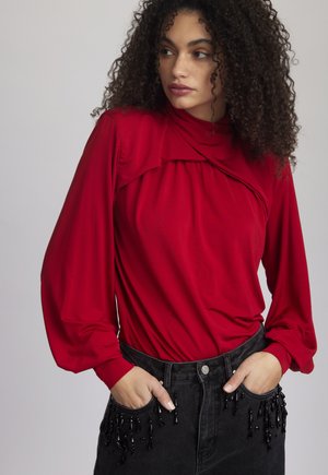 Raudona ilgomis rankovėmis bliusonė su surinkta detalė kaklo srityje ir laisvomis rankovėmis, derinama su juodomis kelnėmis aukšta juosmens linija, papuoštomis karoliukais.