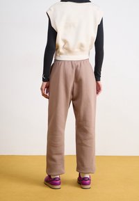 Person wearing beige sweatpants, white sleeveless top over black long-sleeve shirt, and pink Nike sneakers, standing on mustard floor facing away.