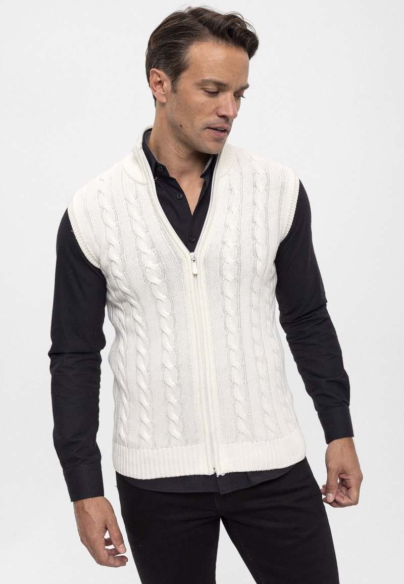 Elfenbeinfarbener Strickpullunder mit Reißverschluss, V-Ausschnitt und geripptem Saum. Wird über einem schwarzen Langarmshirt getragen. Glatte Textur.