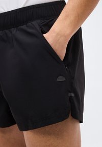 Άτομο που φοράει μαύρο αθλητικό σορτς με το δεξί χέρι στην τσέπη με φερμουάρ στο πλάι, κοντινή λήψη που δείχνει την ελαστική μέση και μια μικρή λεπτομέρεια λογότυπου.