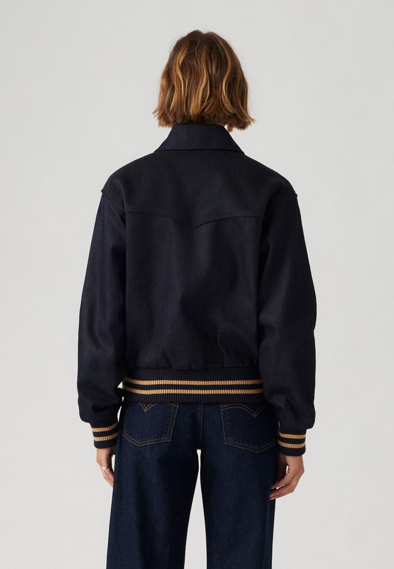 Veste en denim foncé avec une silhouette ajustée, dotée d'une bordure et de poignets côtelés contrastés en marron et crème, avec un col pointu et un détail de dos yoke.