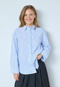 Vaaleansininen napitettava paita, jossa on kirjailtuja kukkakuvioita, pitkät hihat ja pehmeä materiaali, yhdistetty tumman laskostettuun hameeseen.