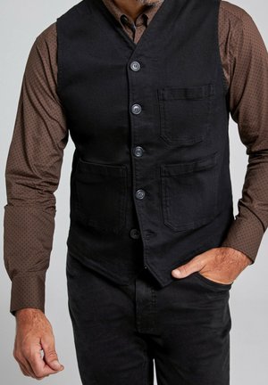 Homme portant un gilet noir boutonné sur une chemise marron à manches longues avec un motif subtil à pois, main dans la poche du pantalon et poils du visage visibles.