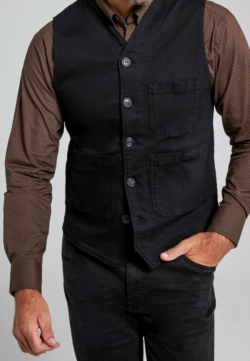 Homme portant un gilet noir boutonné sur une chemise marron à manches longues avec un motif subtil à pois, main dans la poche du pantalon et poils du visage visibles.