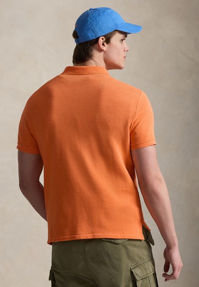 Άνδρας που φορεί πορτοκαλί κοντομάνικο πόλο πουκάμισο, χακί cargo παντελόνι και μπλε καπέλο μπέιζμπολ, φαίνεται από πίσω, κοιτάζοντας προς τα δεξιά.