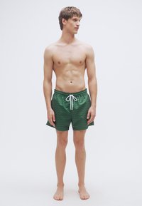 Πράσινα μαγιό με κυματιστό μοτίβο, κορδόνι στη μέση και πλαϊνές τσέπες, παρουσιάζονται σε όρθιο ανδρικό μοντέλο.