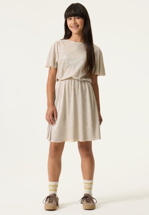 Robe courte beige à finition texturée, manches courtes et taille élastique. Associée à des chaussures marron et des chaussettes rayées crème et jaune.