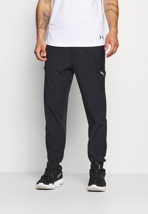 Mężczyzna w czarnych spodniach typu cargo, czarnych trampkach i białej koszulce z krótkim rękawem, stojący na tle jasnego, jednolitego tła.