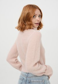 Maglione rosa lavorato a maglia con colletto alto, texture a coste e maniche lunghe. Indossato con jeans in denim chiaro. Aspetto morbido e accogliente.