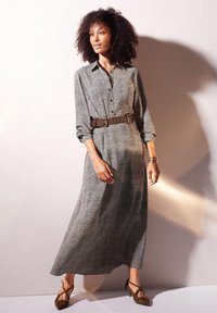 Lange, opgerichte jurk in zwart-wit luipaardprint, getailleerd met een brede bruine riem. Heeft lange mouwen en een vloeiende uitstraling.