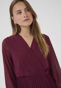 Rochie burgundă cu decolteu în V și mâneci lungi din material transparent, având un tiv plisat. Cercei geometrice argintii adaugă detalii. Fundal simplu.