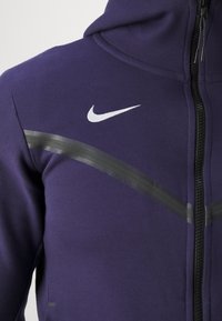 Sweat à capuche zippé violet en tissu doux. Présente un logo Nike blanc et des accents horizontaux noirs sur la poitrine. Comprend une capuche.