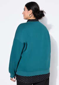 Felpa oversize color teal con polsini e orlo a coste, indossata sopra una camicia nera a fantasia. Tessuto liscio, design minimalista.