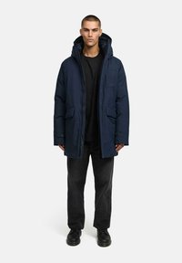 Granatowa pikowana kurtka z kapturem, dwoma kieszeniami z przodu i zapięciem na zamek. Nosi ją w połączeniu z czarną koszulą oraz ciemnymi spodniami i butami.