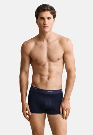 Giovane uomo con capelli corti e scuri che indossa boxer blu navy GANT, in piedi davanti a uno sfondo chiaro e semplice, rivolto verso l'obiettivo.