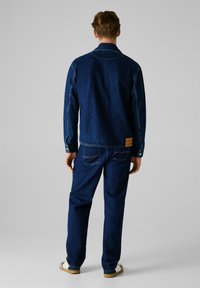 Chaqueta de denim y pantalones en azul oscuro, con cuello estándar, puños de botón y bolsillo trasero de parche. Textura suave, ajuste clásico.