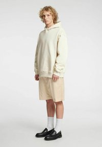 Kremna majica s kapuco in sprednjim žepom, bež kratke hlače, bele nogavice in črne mokasine. Prijeten kroj z razbremenjenim dizajnom.