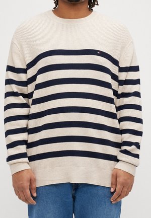 Person iført en beige striktrøje med vandrette sorte striber og blå jeans, stående mod en ensfarvet hvid baggrund.