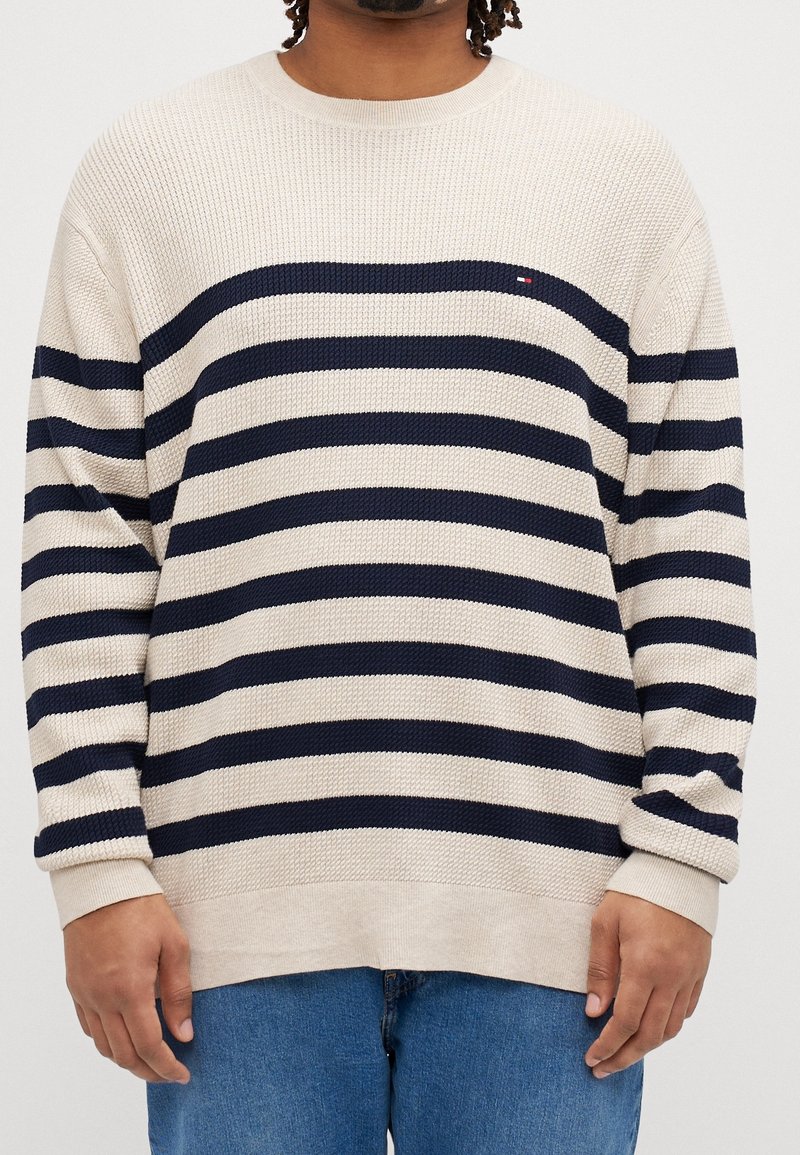 Personne portant un pull en tricot beige avec des rayures noires horizontales et un jean bleu, debout devant un fond blanc uni.